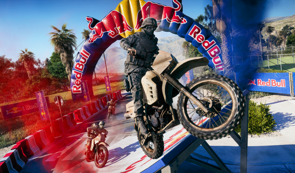 Um soldado em traje tático militar preto empinando uma moto de trilha (Dirt Bike) bege ao passar por baixo de um grande arco inflável da Red Bull em uma pista de corrida ao ar livre, com outros competidores em segundo plano.