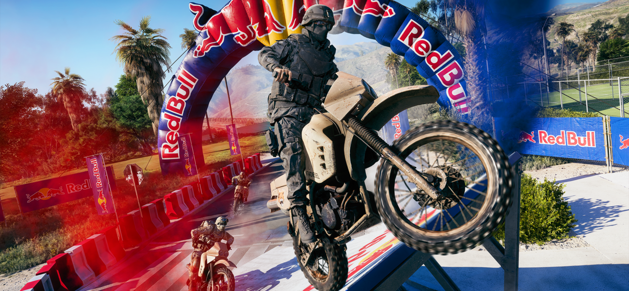 Um soldado em traje tático militar preto empinando uma moto de trilha (Dirt Bike) bege ao passar por baixo de um grande arco inflável da Red Bull em uma pista de corrida ao ar livre, com outros competidores em segundo plano.